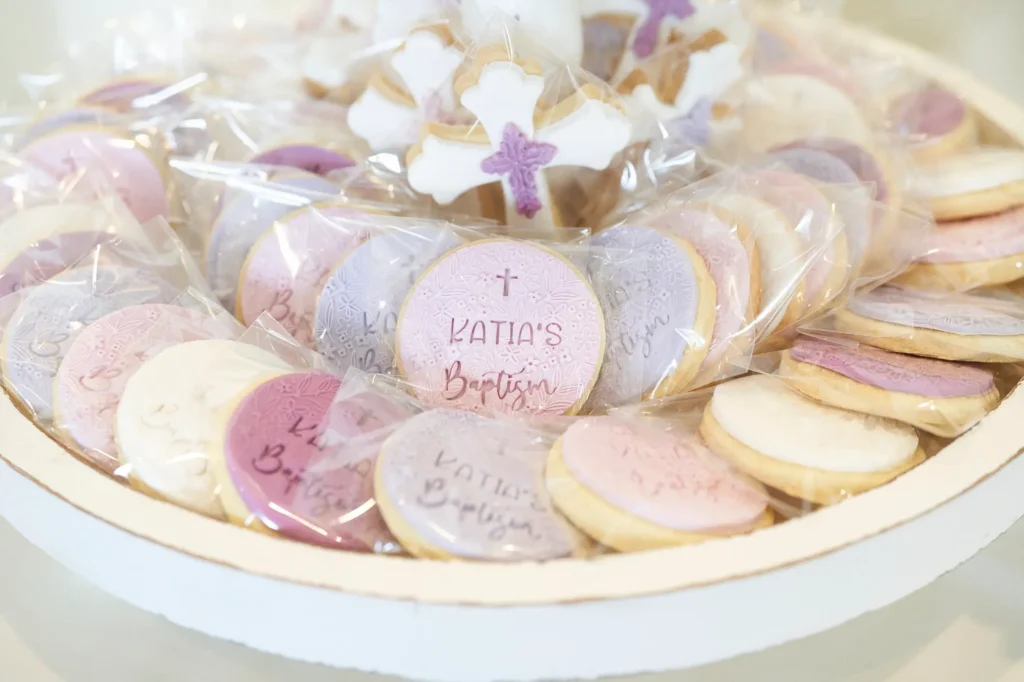 A close-up shot of a white tray filled with individually wrapped sugar cookies made for Katia’s Baptism. The cookies are decorated in pastel colors including pink, lavender, and white, featuring textured icing with elegant script, alongside several cross-shaped cookies with purple accents