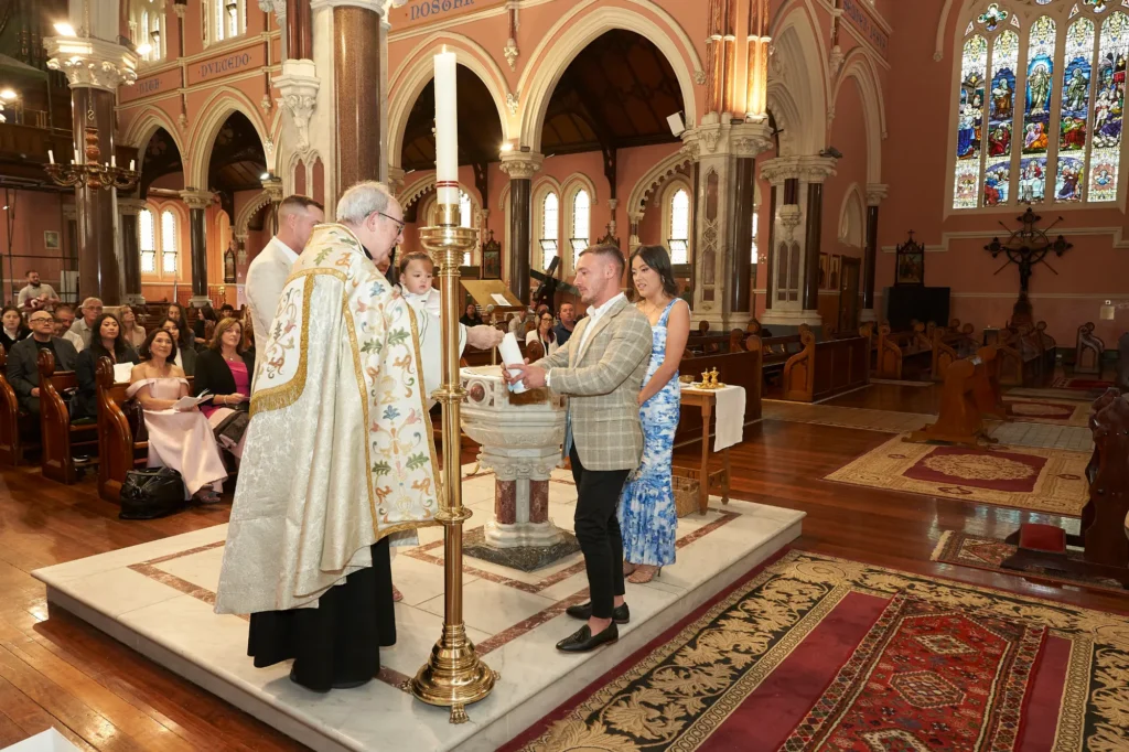 A priest in ceremonial vestments performs a Baptism at a decorative stone font inside St Mary Star of the Sea Church in West Melbourne, Australia. Parents and attendees watch attentively as the ceremony unfolds, with the church’s high arched ceilings, ornate columns, and stained-glass windows creating a solemn and reverent atmosphere that captures the sacred significance of the moment