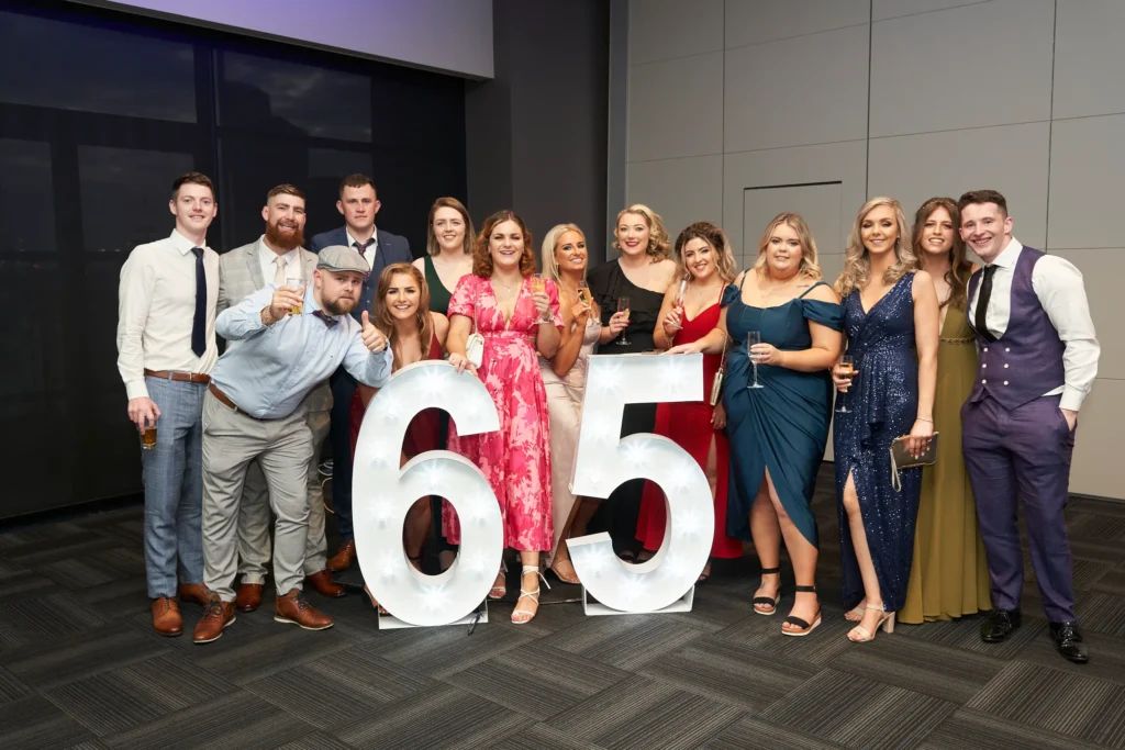 svg%3E A joyful group of approximately fifteen elegantly dressed guests gather around large illuminated “65” numbers, celebrating St. Kevin’s 65th Anniversary Ball. The women wear vibrant formal dresses while the men are in suits and waistcoats. Captured by an event photographer in Melbourne, this image reflects the glamour, excitement, and sense of togetherness of this milestone celebration.