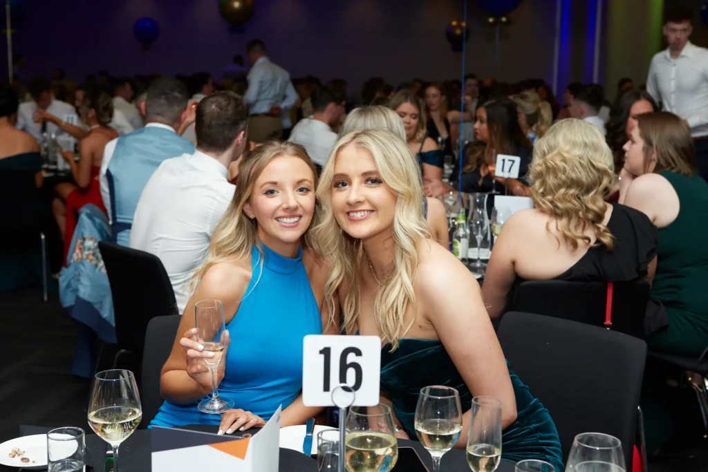 svg%3E Two elegantly dressed women share a joyful moment at St. Kevin’s 65th Anniversary Ball, seated at table 16. One wears a bright blue sleeveless top, and the other a dark green strapless gown, both holding champagne flutes and smiling warmly at the camera. In the background, the ballroom is alive with other guests, festive decorations, and a celebratory atmosphere.