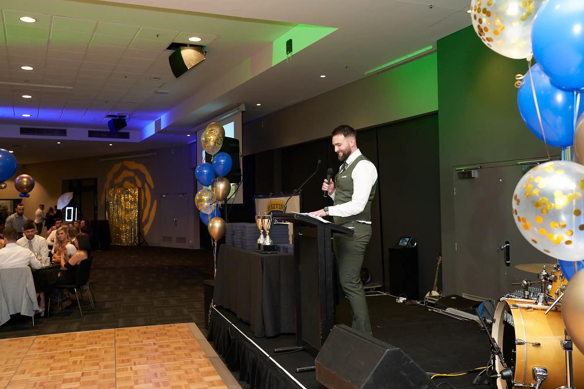 st kevins 65th anniversary ball marvel stadium awards stage A presenter stands at the podium on stage during the Presentation & Awards segment of St. Kevin’s 65th Anniversary Ball at Marvel Stadium. The stage features a displayed award on a draped table, surrounded by festive blue and gold balloons, some with confetti, and green-and-white lighting in the backdrop. In the foreground, elegantly set tables with guests create a formal and celebratory atmosphere, capturing the excitement and significance of this milestone anniversary event.