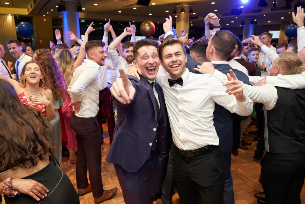 svg%3E Two men in formal attire share a joyful moment on the lively dance floor at St. Kevin’s 65th Anniversary Ball at Marvel Stadium. Behind them, a crowd of guests dances enthusiastically, hands raised in celebration, surrounded by festive decorations and balloons, capturing the vibrant energy and camaraderie of the evening.