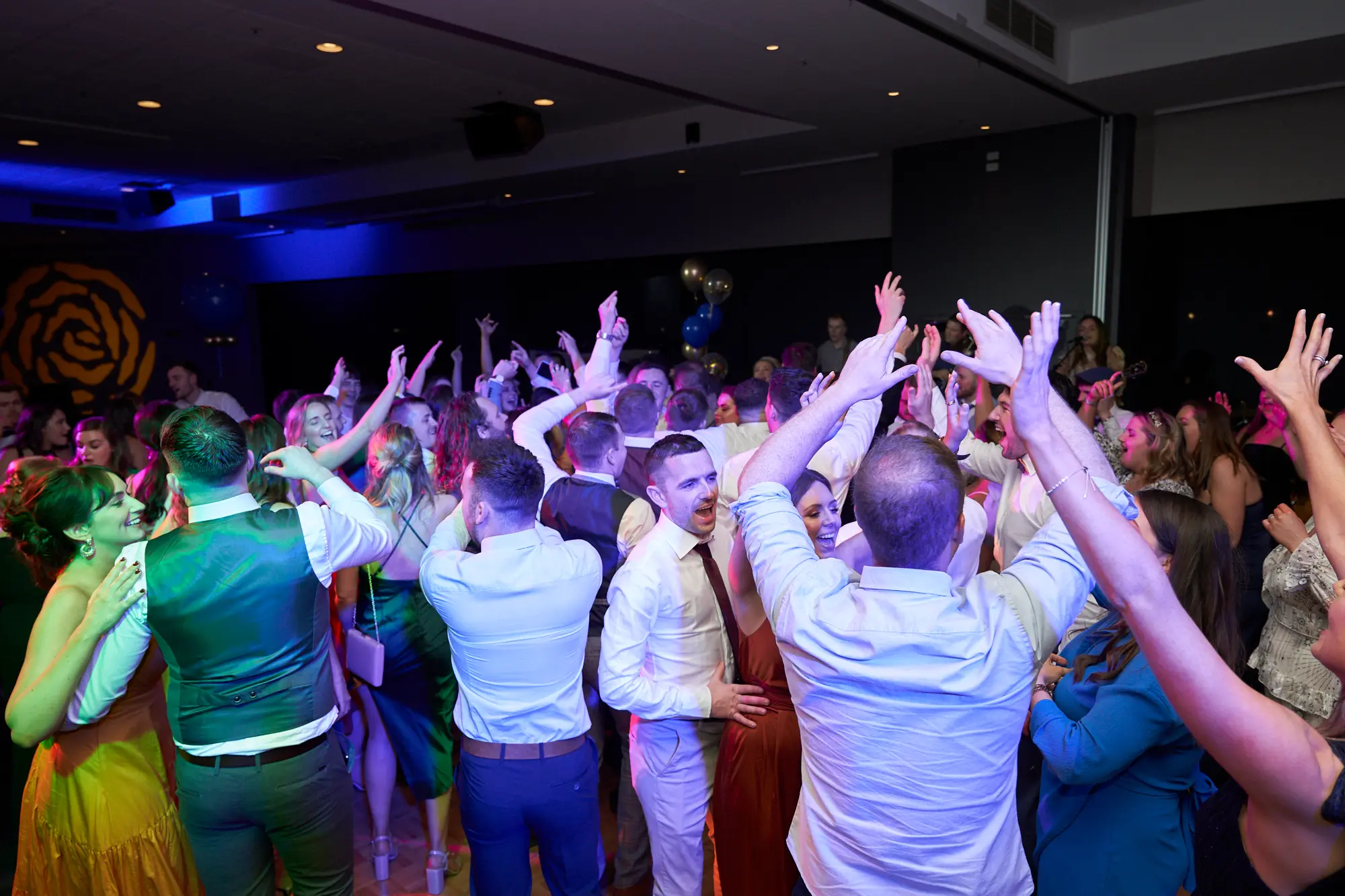 st kevins 65th anniversary ball marvel stadium dance floor energy Energetic dance floor moment at St. Kevin's 65th Anniversary Ball, showing guests in formal attire dancing, raising their arms, and celebrating under vibrant blue and colored lights, capturing the lively and joyful atmosphere of the event.