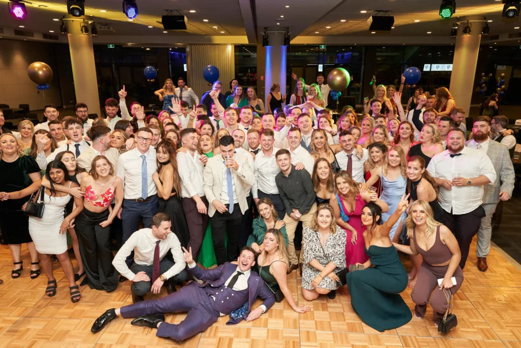 svg%3E A large and diverse group of guests, elegantly dressed in suits and formal gowns, gather together at St. Kevin’s 65th Anniversary Ball at Marvel Stadium. They smile, pose, and interact warmly, conveying joy and camaraderie. The spacious event hall is adorned with festive lighting, balloons, and spotlights, creating a celebratory and lively atmosphere that reflects the excitement and significance of this milestone anniversary celebration.