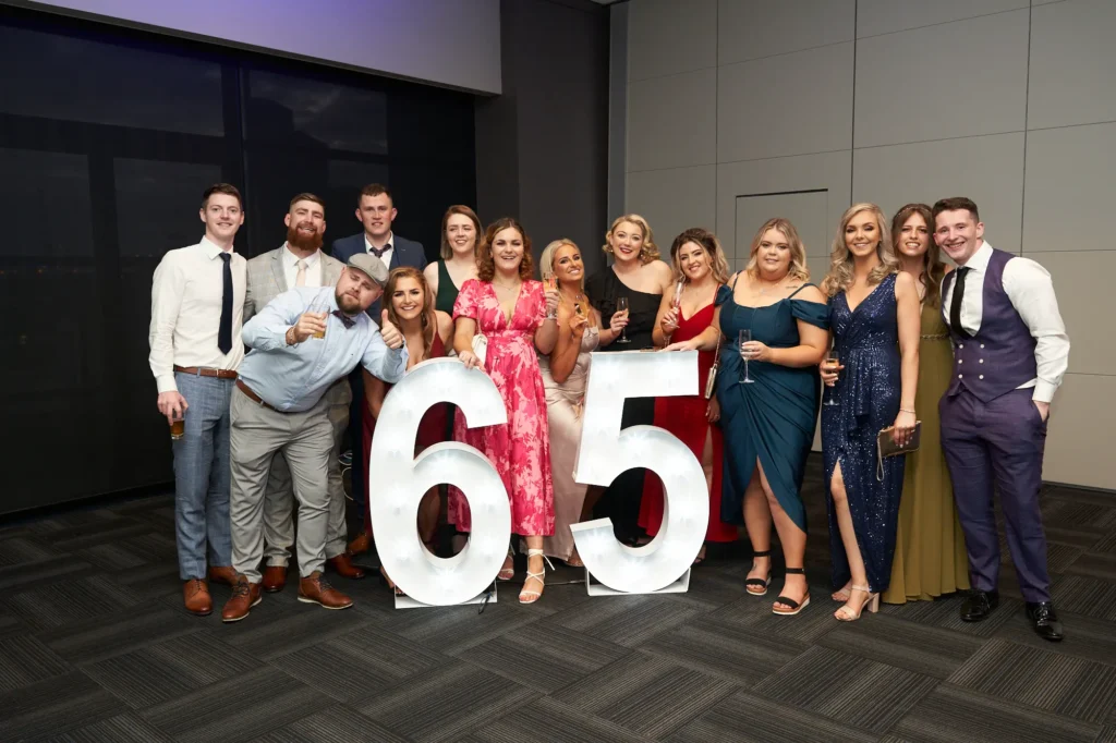 This image captures a lively group photo from St. Kevin’s 65th Anniversary Ball at Marvel Stadium. A diverse group of men and women in elegant formal attire—suits, dresses, and gowns—pose together with joyful smiles. Large illuminated "65" numbers highlight the milestone anniversary, while several guests hold drinks in a celebratory toast. The image conveys a sense of camaraderie, festivity, and shared enjoyment, reflecting the joyous atmosphere of the milestone event.