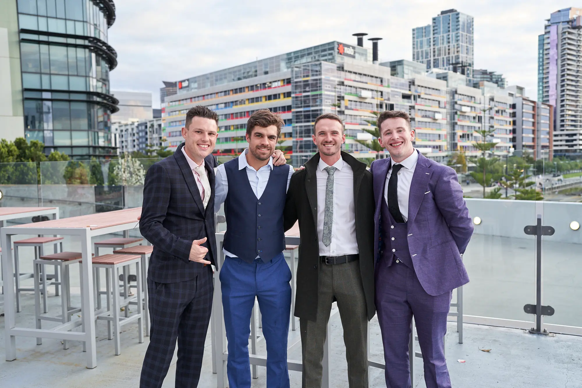 st kevins 65th anniversary ball marvel stadium guest arrivals terrace Four male guests dressed in formal suits, including a checkered suit, three-piece suit, overcoat, and purple suit, pose and smile on an outdoor terrace at Marvel Stadium during St. Kevin’s 65th Anniversary Ball. The contemporary urban skyline in the background enhances the celebratory and elegant atmosphere as guests arrive for the milestone event
