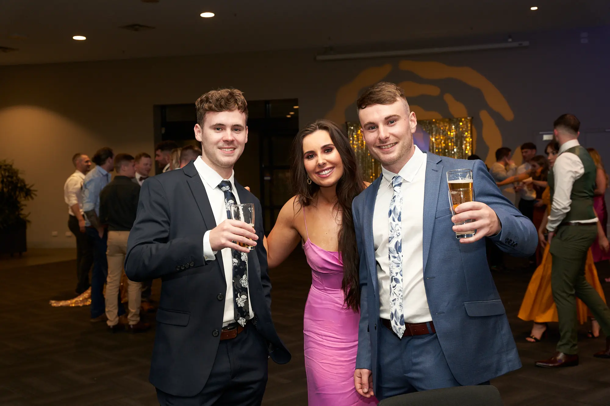 st kevins 65th anniversary ball marvel stadium guest celebration candid his candid image from St. Kevin’s 65th Anniversary Ball captures three guests sharing a joyful moment. Two men in suits and a woman in a pink dress smile warmly at the camera while holding drinks, embodying the celebratory spirit of the evening. Behind them, the ballroom buzzes with activity, with other attendees mingling and enjoying the festivities. Subtle hints of decor in the background and the lively interactions among guests convey the vibrant, social atmosphere of this milestone anniversary celebration at Marvel Stadium.