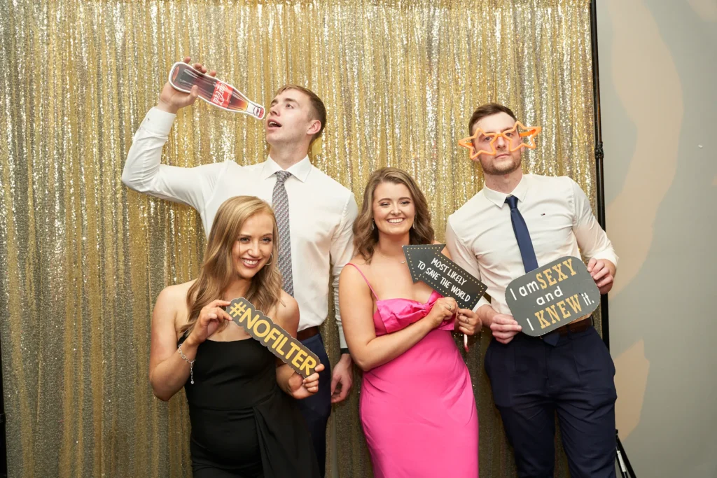 svg%3E Four guests strike playful poses in a photobooth at St. Kevin’s 65th Anniversary Ball at Marvel Stadium. Two men in suits hold humorous props, including oversized glasses and a novelty beverage bottle, while two women in elegant pink and black dresses hold fun signs like “MOST LIKELY TO SAVE THE WORLD” and “#NOFILTER.” The shimmering gold backdrop adds to the lighthearted and celebratory atmosphere of the evening.