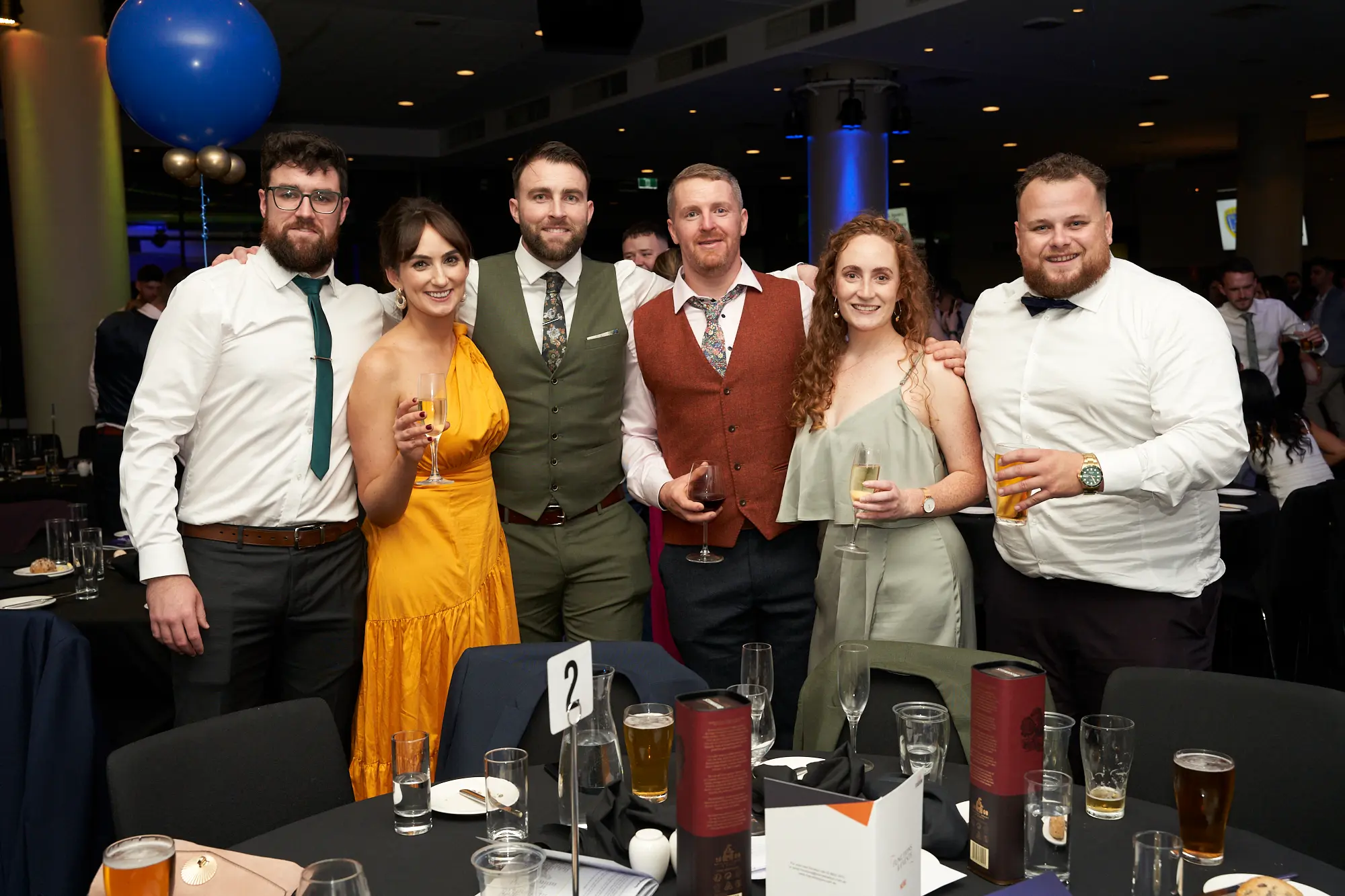 st kevins 65th anniversary ball marvel stadium table2 group Six elegantly dressed guests, three men and three women, gather around table 2 during St. Kevin’s 65th Anniversary Ball at Marvel Stadium. The women wear vibrant yellow and muted green gowns, while the men are in shirts, ties, and a vest. Drinks are raised and smiles abound, with a large blue balloon in the background, capturing the festive atmosphere and shared connections of this milestone celebration