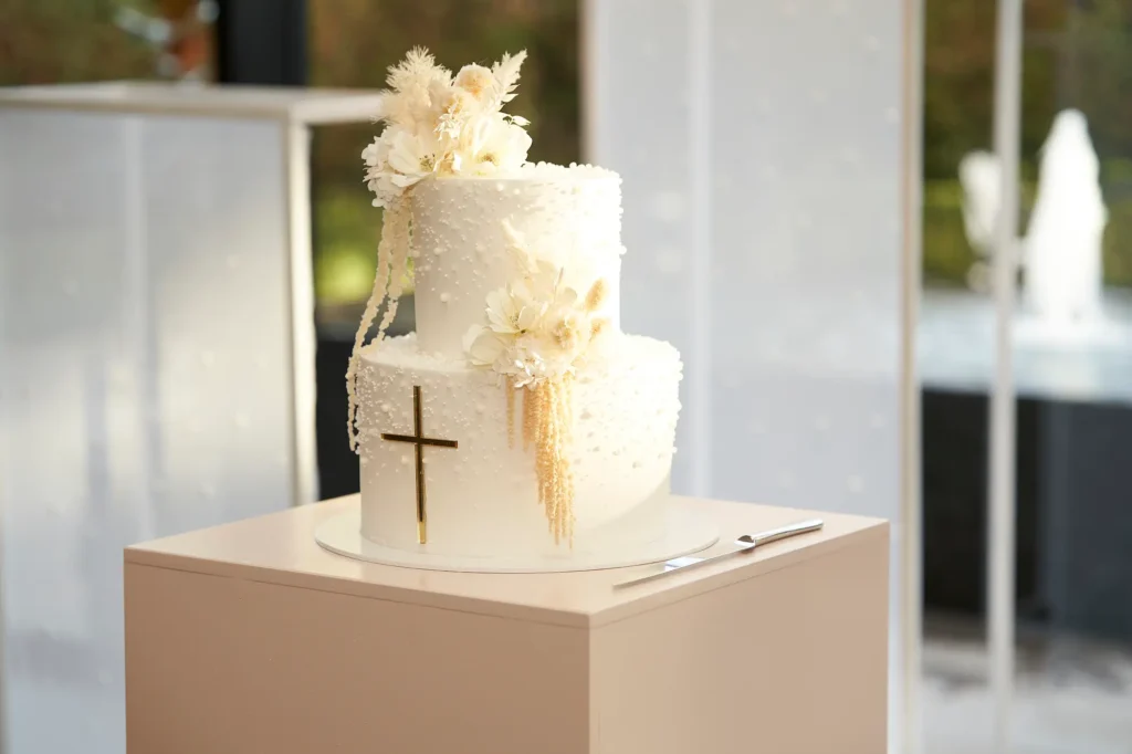 An elegant two-tier white cake decorated with delicate white and cream flowers and pampas grass, featuring a gold cross on the bottom tier. The cake is displayed on a pedestal during a reception, highlighting the meaningful details that make the celebration memorable