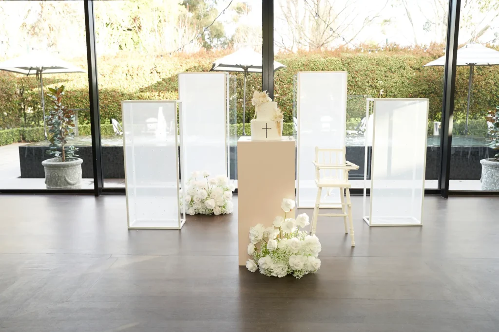 A white-tiered cake topped with a cross and adorned with delicate white flowers sits on a tall beige pedestal in a bright room with large windows overlooking a garden. Flanking the pedestal are arrangements of white roses and baby's breath, a high chair, and transparent display cases, capturing the elegant and celebratory details of a Baptism event