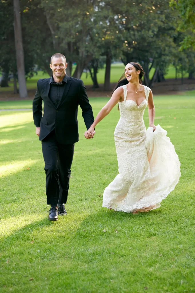 A joyful candid moment of a newly married couple running hand in hand through the lush green lawns of Treasury Gardens in Melbourne. The groom, dressed in a black suit and bow tie, smiles brightly toward the camera, while the bride, in a lace white gown, lifts her dress as she laughs and looks to the side. Sunlight filters through the trees, casting soft shadows across the grass and creating a lively, carefree atmosphere. This image beautifully captures the playful energy and pure happiness of the couple as they celebrate their wedding day in a serene park setting.