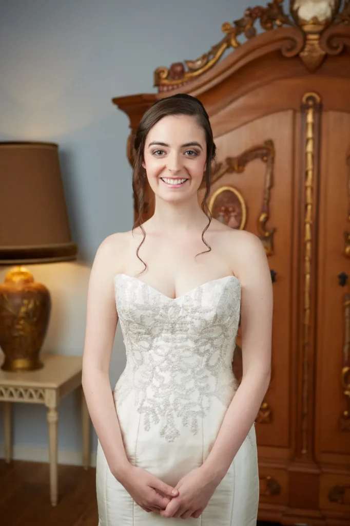 A serene and elegant bridal portrait showing the bride standing gracefully in a classic, well-decorated room. She faces the camera with a bright, relaxed smile and her hands gently clasped in front of her. Her strapless wedding gown features an intricately beaded bodice with a sweetheart neckline and a sleek satin skirt. Her dark hair is styled in a timeless updo with soft curls framing her face. Behind her, antique furniture, including a carved wooden armoire and a gold-based table lamp, enhances the room’s vintage charm. This Melbourne wedding getting-ready moment radiates poise, happiness, and timeless sophistication.