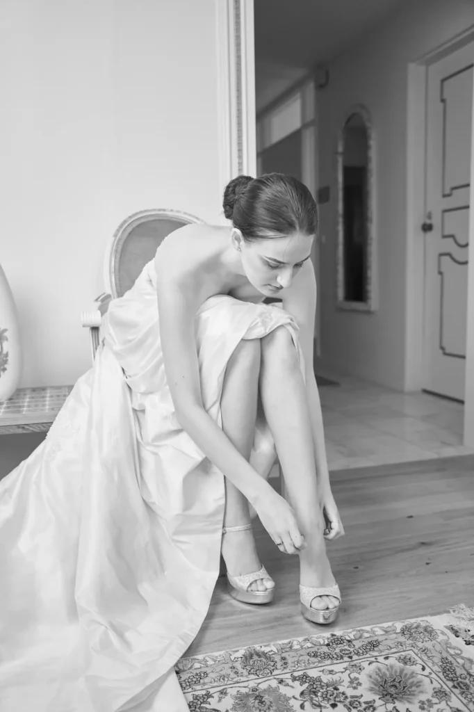 A quiet and intimate black-and-white portrait of a bride preparing for her wedding day. Seated on a formal chair, she focuses on adjusting the ankle strap of her high-heeled shoe, her flowing strapless gown pooling gracefully around her. The light-colored wooden floor and a decorative patterned rug add elegance to the scene, while soft natural light illuminates her and the surrounding room. In the background, an open doorway reveals a hallway with an ornate mirror, hinting at the bridal suite’s refined details. The photograph captures a serene, personal moment of anticipation and preparation before the ceremony.