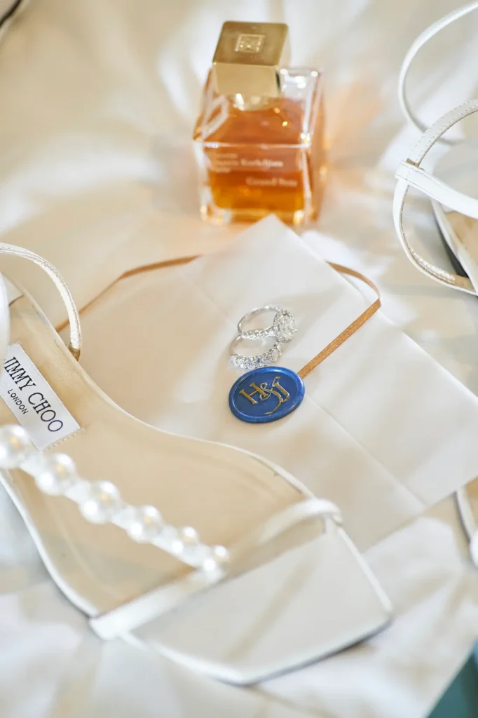 A beautifully styled flat lay of the bride’s wedding day accessories, captured on a soft white textured surface. At the forefront are elegant white Jimmy Choo high-heeled sandals, one featuring a delicate pearl strap detail. In the center, two silver rings — a diamond engagement ring and a wedding band — rest atop a white cloth and a blue wax seal embossed with gold “H&J” initials. Behind them, a square perfume bottle with a gold cap and amber liquid adds a touch of warmth. This refined composition highlights the timeless elegance of a bride getting ready for her wedding day.