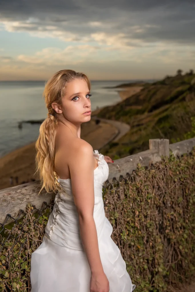 A bride stands beside a rustic wooden fence overlooking the ocean at sunset, her strapless gown glowing softly in the warm evening light. Her blonde hair is styled in a loose side ponytail as she leans gently on the fence, glancing back toward the camera with a poised, contemplative expression. The pastel hues of the sky blend beautifully with the rolling grassy cliffs and the distant headland, creating a serene coastal atmosphere. Bathed in golden light, the portrait evokes a feeling of quiet strength and timeless romance, capturing the elegance of the bride against the breathtaking seaside landscape.