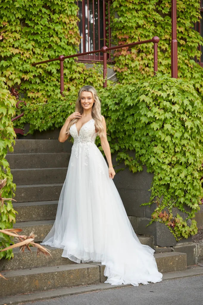 A radiant bride stands gracefully on the stone stairs of Victoria Barracks, surrounded by a lush ivy-covered wall that creates a romantic, storybook setting. Her white wedding gown flows beautifully around her feet as she turns slightly toward the camera with a warm, gentle smile. The soft natural light enhances the serene atmosphere, highlighting the delicate textures of her dress and the vibrant greenery around her. Captured in Melbourne, this wedding portrait at Victoria Barracks blends timeless elegance with natural beauty, perfectly reflecting the calm joy and grace of the bride on her special day.
