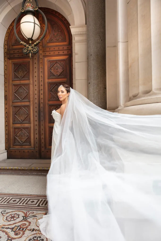 A stunning bridal portrait captured outside the Parliament House of Victoria, showcasing a bride standing before a grand wooden door framed by tall, fluted columns. Her elegant white gown and flowing veil billow gracefully behind her as she looks over her shoulder toward the camera with poise and confidence. The intricate architecture and mosaic floor add depth and sophistication, creating a perfect setting for this timeless Melbourne wedding photography moment. Soft, glowing light enhances the scene’s elegance, highlighting the textures of her dress and the ornate details of the Parliament House, blending romance, architecture, and artistry beautifully.
