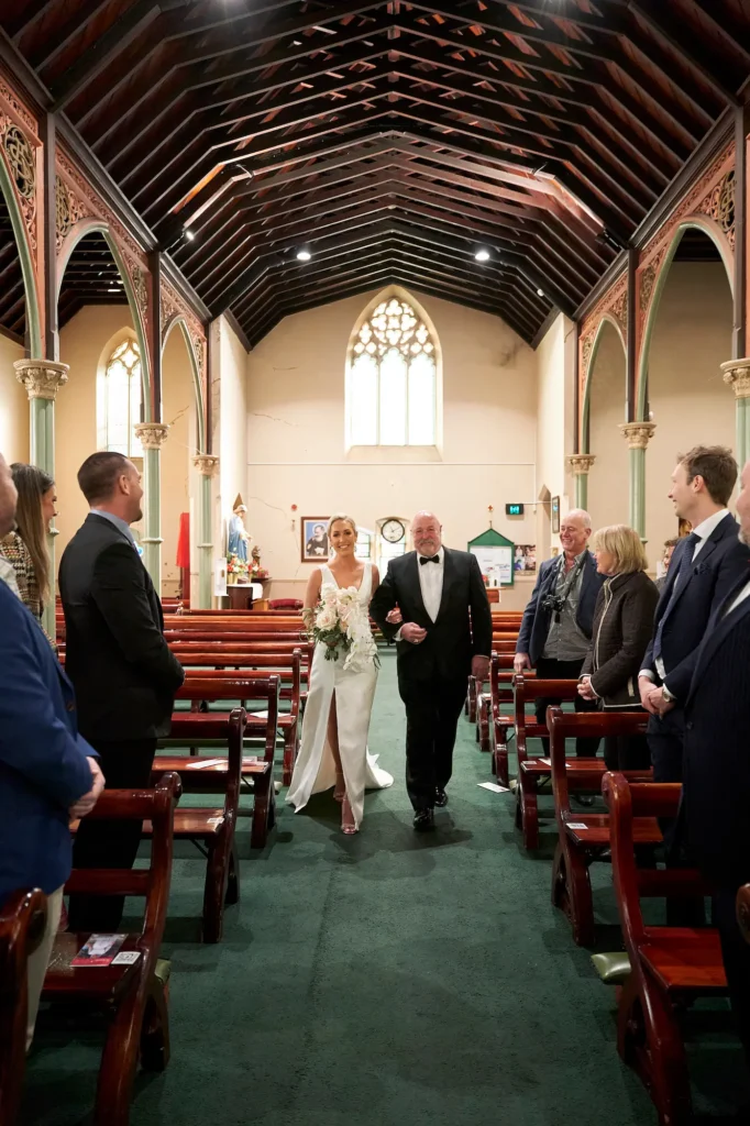This heartwarming wedding photograph captures a bride walking down the aisle at St. Augustine Parish in Melbourne, escorted by her father. Smiling joyfully, she makes her way toward the altar as family and friends look on with love and excitement. The church’s elegant wooden pews, green-carpeted aisle, and tall arched windows create a warm, intimate atmosphere. Sunlight filters gently through the stained glass, illuminating the bride’s radiant expression. This emotional moment, beautifully documented through Melbourne wedding photography, reflects the joy, love, and timeless beauty of a heartfelt wedding ceremony.