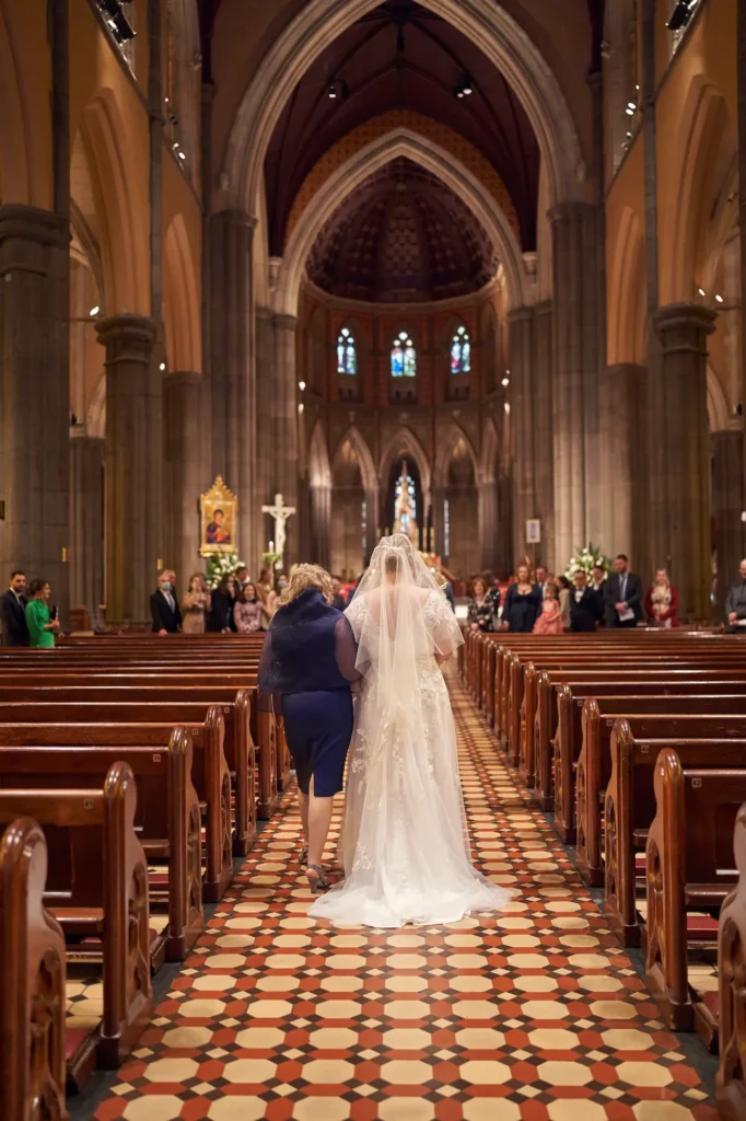 This emotional wedding photograph captures a bride walking down the aisle at St Patrick’s Cathedral in Melbourne, accompanied by her mother. The cathedral’s grand Gothic arches, patterned tiled floor, and soft light filtering through stained-glass windows create an awe-inspiring atmosphere. Guests seated along the pews watch in quiet reverence as the bride’s long veil trails behind her. This timeless moment, beautifully captured through Melbourne wedding photography, reflects love, anticipation, and the sacred beauty of a wedding ceremony in one of the city’s most iconic churches.