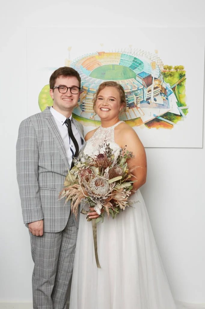 A joyful indoor portrait capturing a heartfelt moment between a bride and her brother on the wedding day. They stand side by side, smiling brightly at the camera. The bride holds a large, rustic bouquet made of protea, eucalyptus, and dried grasses tied with a soft ribbon, while her brother stands with his hands clasped in front of him. She wears a sleeveless white gown with a lace-detailed halter neckline and a flowing skirt. Behind them, a white wall and colorful framed artwork depicting a sports stadium add a modern, personal touch to this warm family moment.