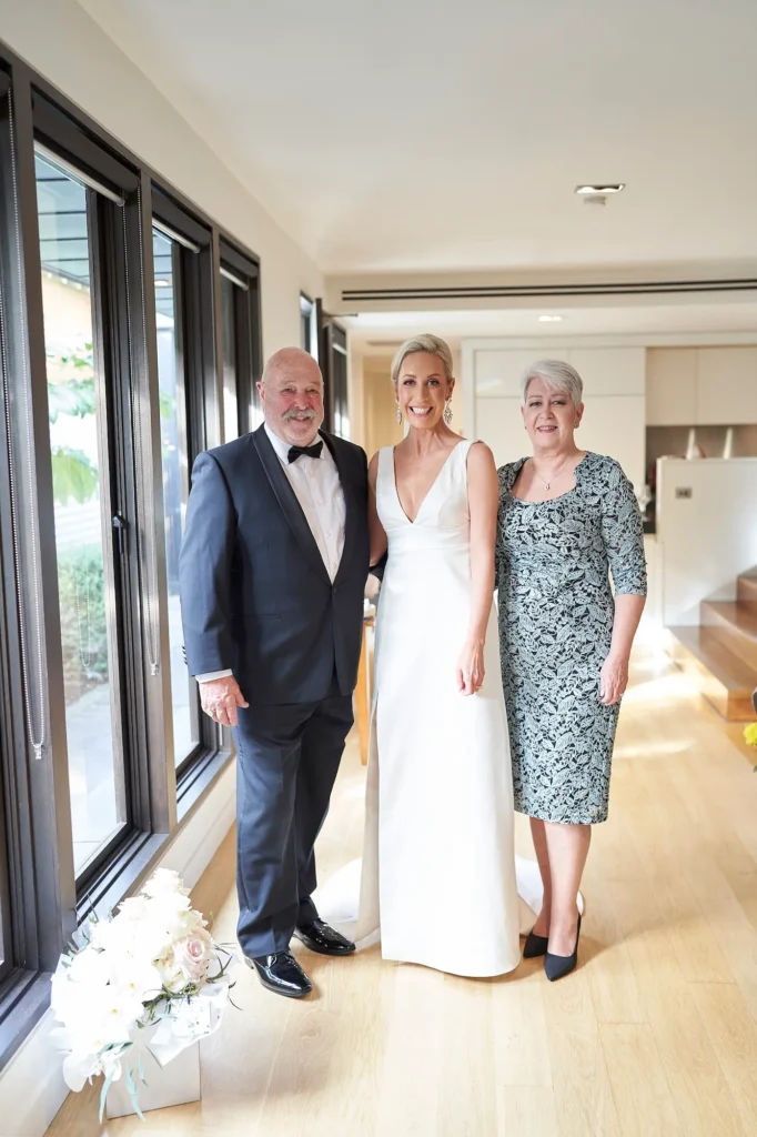 A joyful indoor portrait capturing the bride with her parents during the getting ready phase of her wedding day. Standing in the center, the bride smiles brightly at the camera while her father, on her left, wraps an arm around her shoulder, and her mother, on her right, links arms with her lovingly. The light-filled modern room features floor-to-ceiling windows, wooden floors, and soft natural light that enhances the warm, celebratory mood. The bride’s elegant white gown with a deep V-neckline and flowing skirt adds timeless grace to this heartfelt family moment before the ceremony.