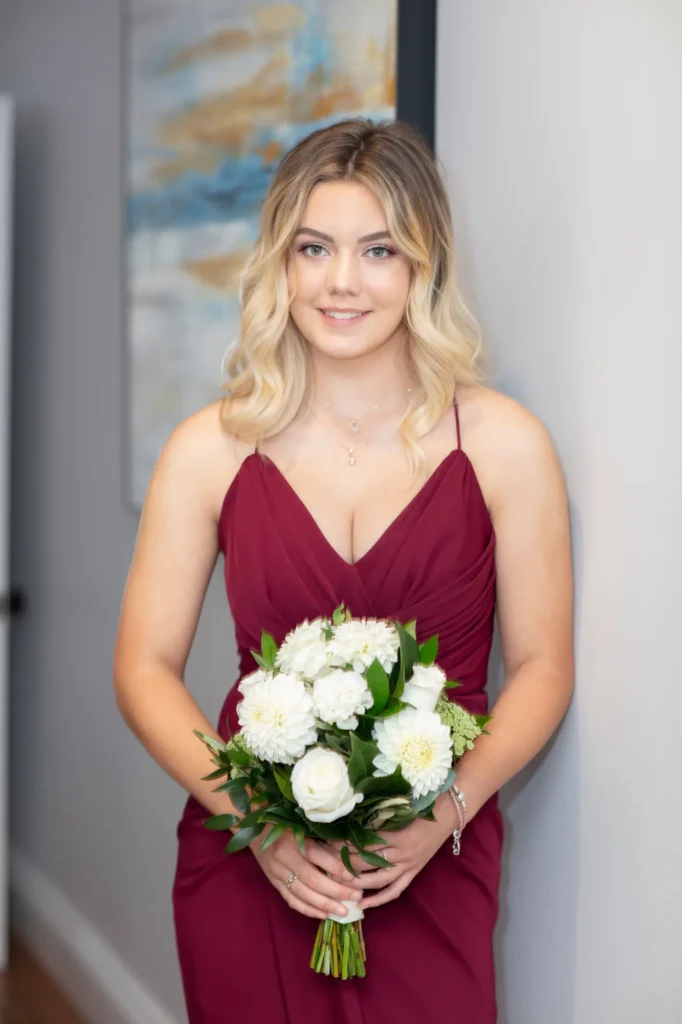 A calm and elegant indoor portrait of a bridesmaid standing against a light gray wall, holding a bouquet of white flowers with both hands at her waist. She faces the camera with a gentle, relaxed smile, exuding warmth and grace. Her sleeveless burgundy dress features thin spaghetti straps and a soft V-neckline, the smooth fabric catching the light with a subtle sheen. To her left, a framed abstract artwork in blue and brown tones adds a touch of color to the minimalist setting, creating a refined and balanced composition that highlights the beauty of this bridal moment.