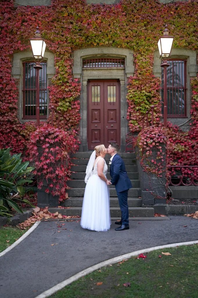 This wedding photograph captures a romantic and intimate moment between the bride and groom, Eden and Corey, at the historic Victoria Barracks in Melbourne, Australia. The couple is standing on a wide set of worn stone steps, sharing a loving kiss, with the groom’s arm around the bride’s waist and his other hand gently holding her back. The bride holds her bouquet with both hands as they focus entirely on each other, creating a sense of deep affection and joy. The background features the bluestone facade of Victoria Barracks, almost entirely covered in vibrant red and crimson ivy, with a large dark red double door and scattered autumn leaves on the steps. This setting provides a picturesque and seasonal scene, ideal for wedding photography Victoria Barracks and Melbourne.