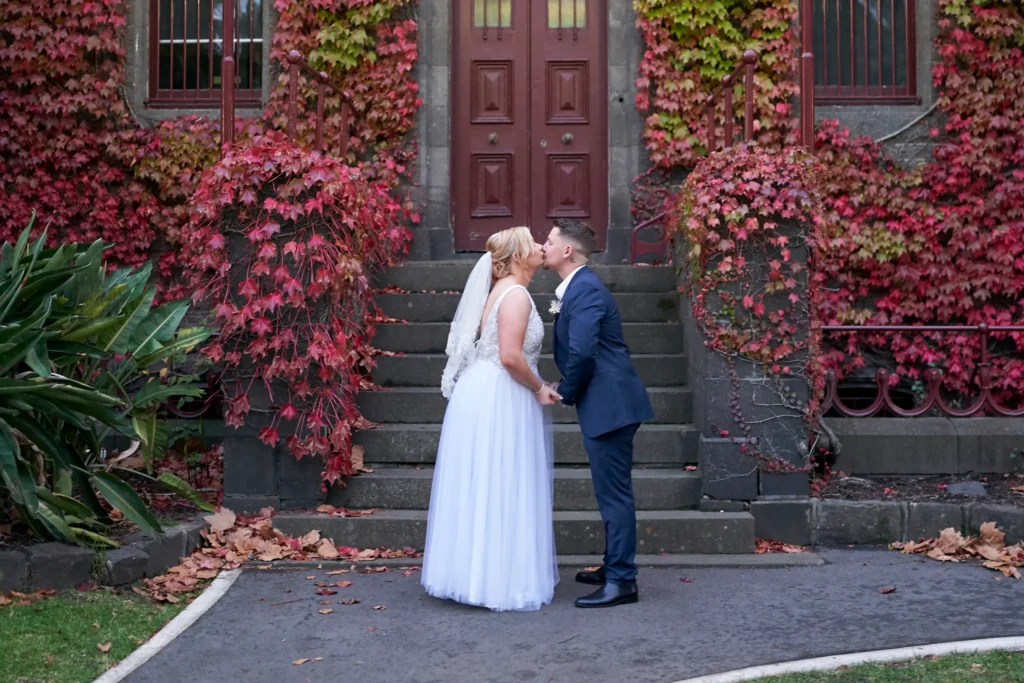 Eden and Corey share an intimate kiss while holding hands at Melbourne’s historic Victoria Barracks, surrounded by vibrant red and orange autumn ivy. The couple’s wedding attire contrasts beautifully with the dark bluestone walls and grand Renaissance Revival architecture. The stone steps and ornate facade create a timeless and romantic backdrop, highlighting the bride’s flowing gown and the celebratory mood. This iconic location, renowned for its historic charm and picturesque autumn scenery, perfectly captures the elegance, romance, and emotional significance of their wedding portraits in Melbourne.