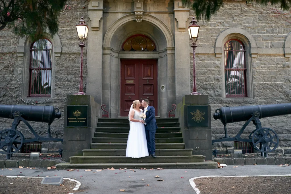 Eden and Corey pose elegantly at Melbourne’s historic Victoria Barracks, surrounded by the imposing 19th-century bluestone building and Renaissance Revival architecture. The couple’s formal wedding attire contrasts beautifully with the rustic, historic setting, enhanced by colorful ivy and two historical cannons flanking the stairs. The iconic steps provide a classic backdrop for the bride’s flowing gown, creating a romantic and timeless wedding portrait that captures both their love and the military and historical charm of Victoria Barracks, a favored location for Melbourne wedding photography.