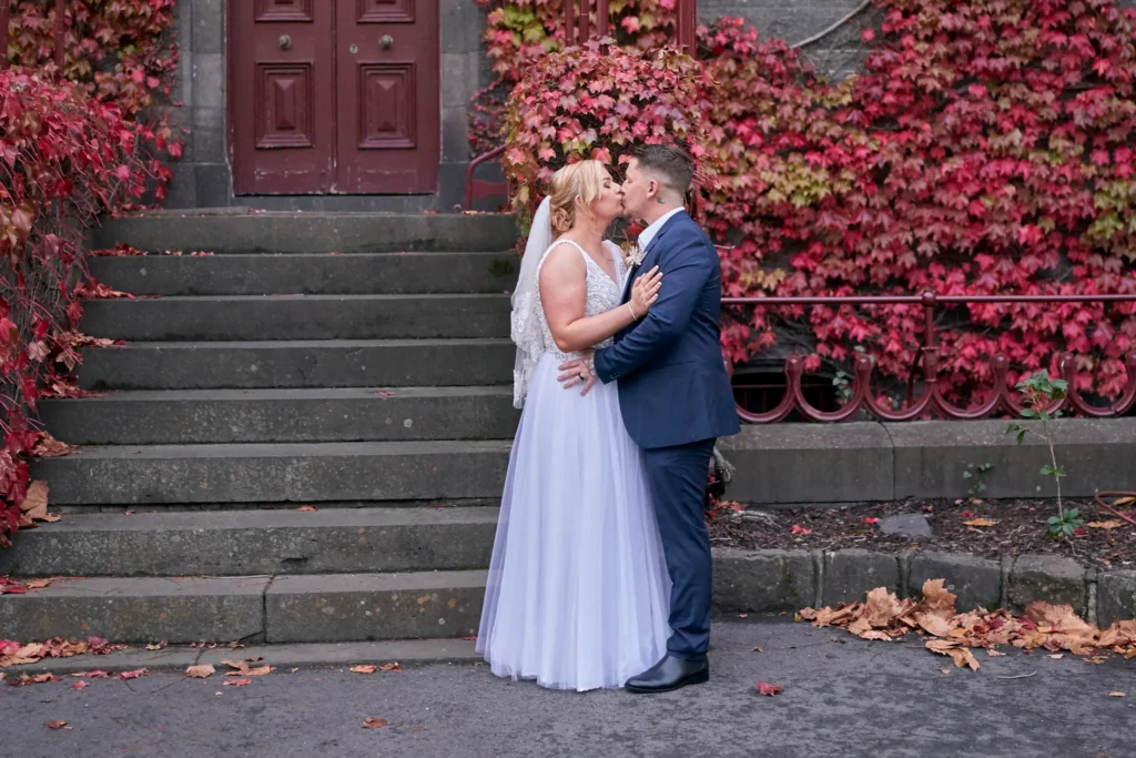 eden corey victoria barracks wedding melbourne couple kiss autumn Eden and Corey share a tender kiss while holding hands in front of the historic Victoria Barracks in Melbourne, surrounded by vibrant red and orange autumn foliage. The bride’s elegant white gown and the groom’s classic blue suit contrast beautifully with the gothic-style architecture, bluestone walls, and iconic stone gate. This romantic setting, known for its timeless charm and seasonal beauty, creates the perfect backdrop for their wedding portrait, capturing both their love and the enchanting atmosphere of Victoria Barracks, a favorite location for Melbourne wedding photography.