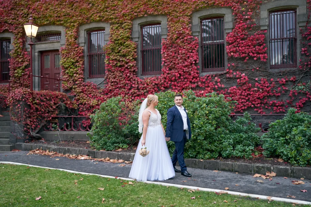 Eden and Corey walk hand in hand at Melbourne’s historic Victoria Barracks, sharing smiles and joy in a candid moment that captures the beginning of their life together. The couple’s wedding attire contrasts beautifully with the 19th-century bluestone walls and Renaissance Revival architecture. Autumn ivy in golden and crimson shades covers the facade, creating a warm and romantic atmosphere. This picturesque location, known for its timeless charm and seasonal beauty, provides a stunning backdrop for wedding photography, highlighting both the couple’s love and the elegance of Victoria Barracks in Melbourne.