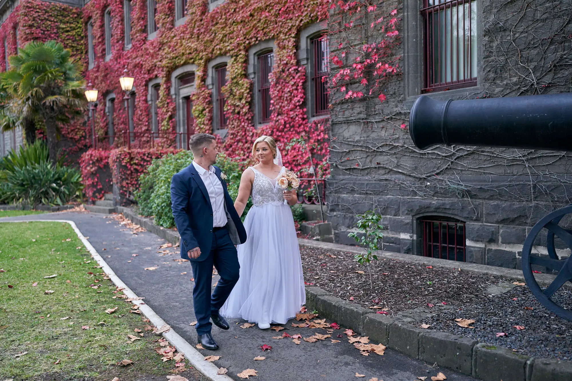 eden corey wedding photography victoria barracks couple walk Eden and Corey share a romantic and intimate moment walking hand-in-hand at the historic Victoria Barracks in Melbourne, Australia. Smiling at each other, the couple radiates love and joy on their wedding day. The Renaissance Revival bluestone building provides a striking backdrop, with walls covered in vibrant red and pink autumn ivy. A black cannon, cobblestone path, and vintage street lamps add to the timeless and historic atmosphere. Scattered autumn leaves enhance the warm and romantic feeling of the scene. This beautifully composed image captures a classic bride and groom portrait at Victoria Barracks, making it an ideal example of elegant wedding photography in Melbourne, full of emotion, history, and seasonal charm.