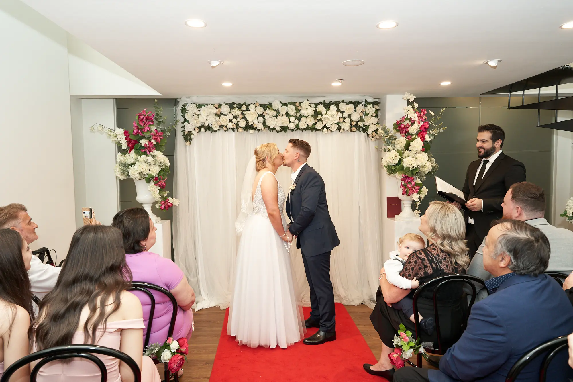 eden corey wedding registry office st kilda road towers ceremony Eden and Corey share a romantic kiss at the St Kilda Road Towers Registry Office during their wedding ceremony, standing on a red carpet with a decorated white backdrop featuring white roses, green foliage, and crimson accents. The bride wears a sleeveless gown with a long veil, and the groom is in a dark blue suit with a black bow tie. Guests, including a woman holding a baby, watch the heartfelt moment, while the officiant smiles nearby, creating an intimate and joyful atmosphere in the modern, elegantly decorated ceremony room.