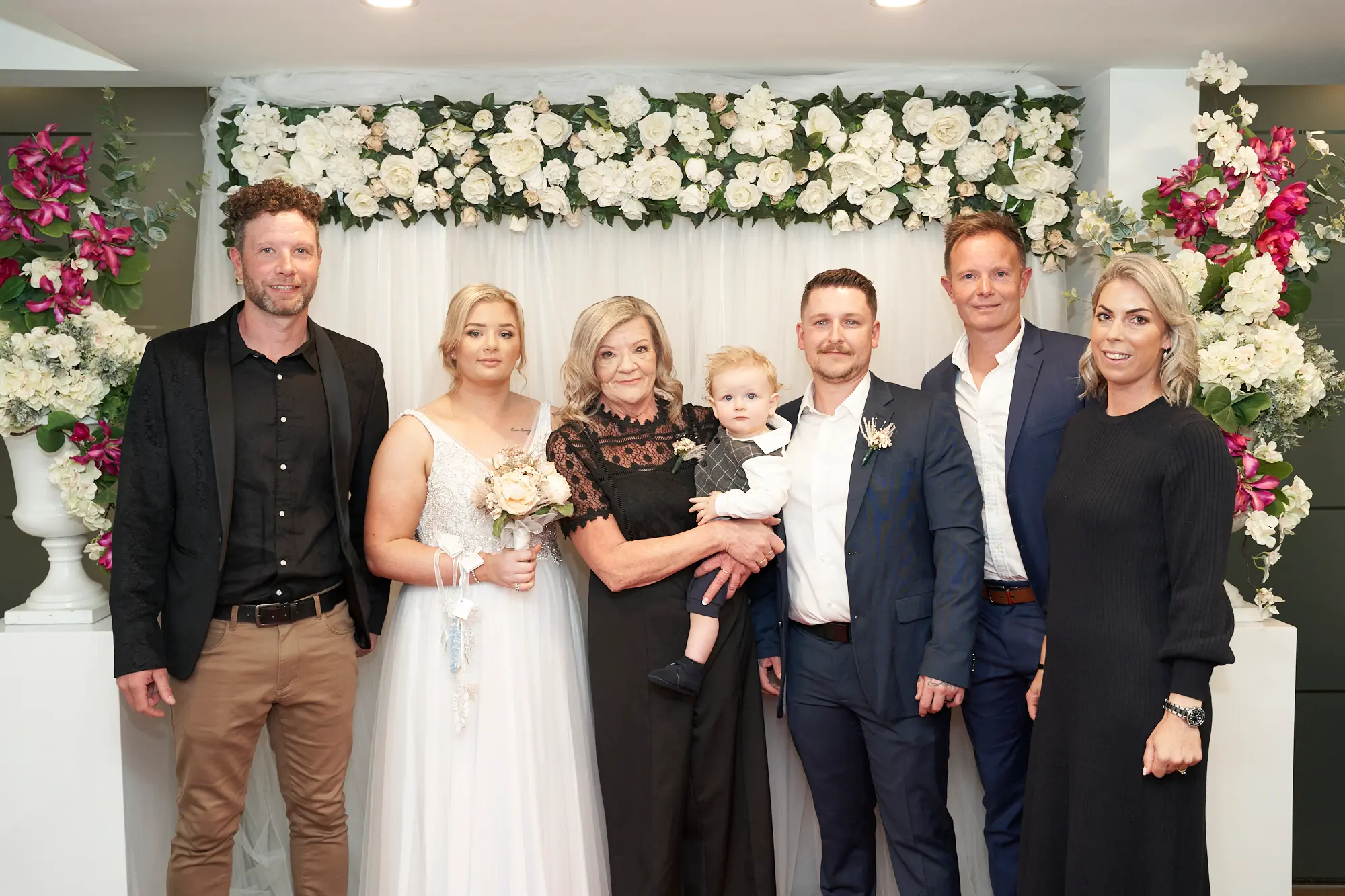 eden corey wedding st kilda road towers family portrait Eden and Corey pose with their family at the St Kilda Road Towers Registry Office, standing in front of a white floral backdrop adorned with white roses and greenery, flanked by large floral arrangements of pink, white, and fuchsia flowers. Eden wears a sleeveless white gown and holds a bouquet of light pink and white flowers, while Corey is in a dark blue suit, holding their young child in a grey suit. Family members stand on either side, smiling warmly, creating a joyful and celebratory group portrait that captures the love and happiness of the wedding day.