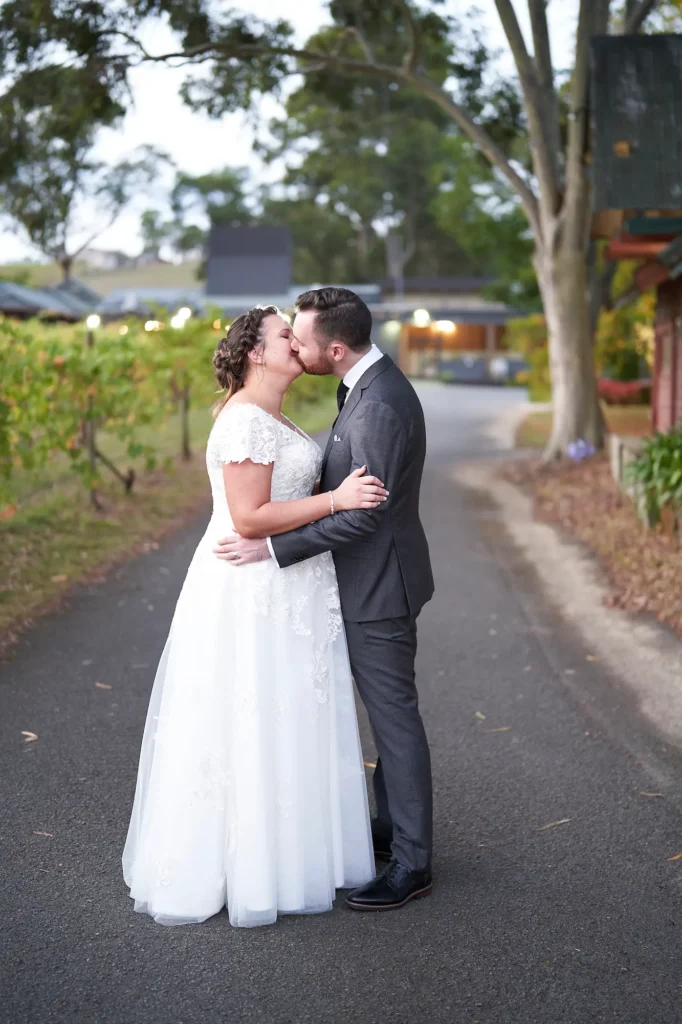 A newly married couple shares a tender kiss along a paved path at Fergusson Winery in the Yarra Valley. The groom, dressed in a dark grey suit, holds his bride close as her lace wedding gown flows softly in the golden evening light. Rows of grapevines stretch into the distance beside a rustic building adorned with glowing string lights, creating a warm, romantic glow. The twilight sky casts a serene blue tone over the scene, blending elegance and intimacy. This Yarra Valley wedding portrait perfectly captures the charm of vineyard romance and the timeless beauty of wedding photography in Fergusson Winery.