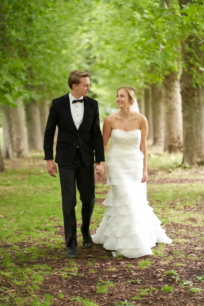 This romantic outdoor wedding portrait captures a bride and groom walking hand-in-hand along a tree-lined path at Fitzroy Gardens in Melbourne. The bride wears a strapless, ruffled white gown with a short veil, holding a small bouquet, while the groom is in a classic black tuxedo with a bow tie. Sunlight filters softly through the lush green canopy above, creating a dappled, serene atmosphere. Their loving glances and relaxed stride convey intimacy and joy, perfectly reflecting the elegance and charm of a Fitzroy Gardens wedding, with a peaceful, natural backdrop framing this tender, celebratory moment.