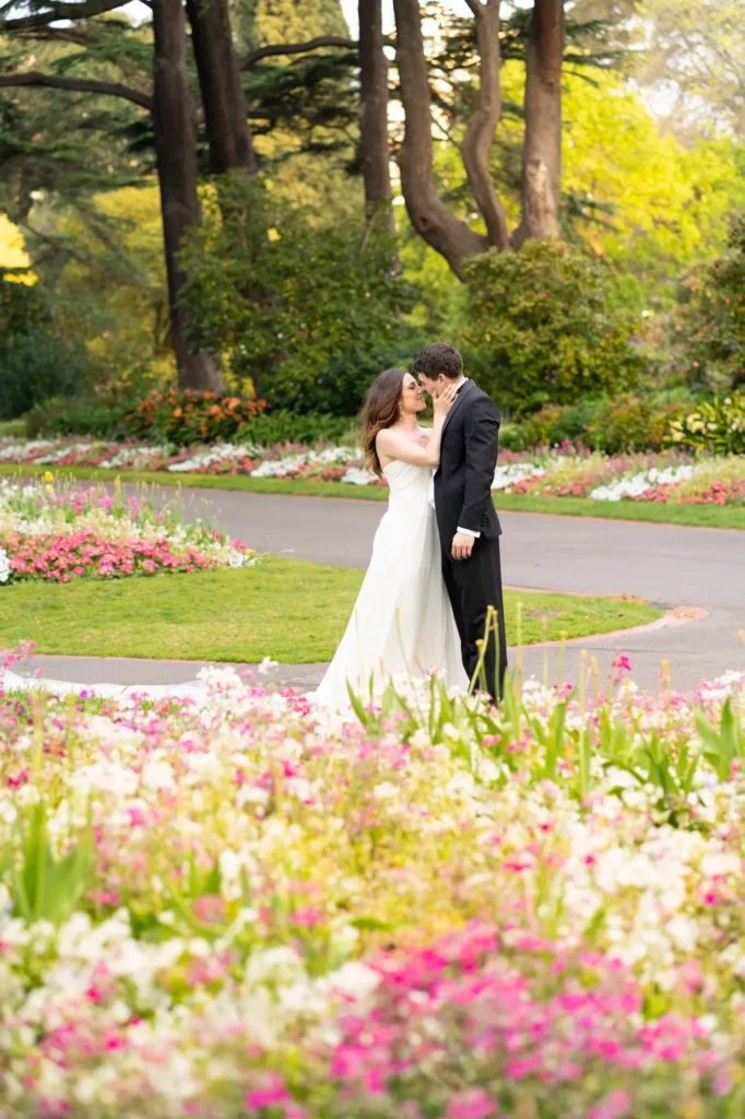 A bride and groom share an intimate moment on a winding path surrounded by vibrant wildflowers in Fitzroy Gardens. The groom, dressed in a classic black tuxedo, gently holds his bride as she looks up at him with a soft smile, their foreheads touching in a tender embrace. Sunlight filters through the trees, illuminating the colorful blooms and casting a warm glow over the scene. The lush greenery and romantic atmosphere capture the essence of love and connection, making this portrait a timeless example of wedding photography in Fitzroy Gardens — elegant, heartfelt, and beautifully natural.