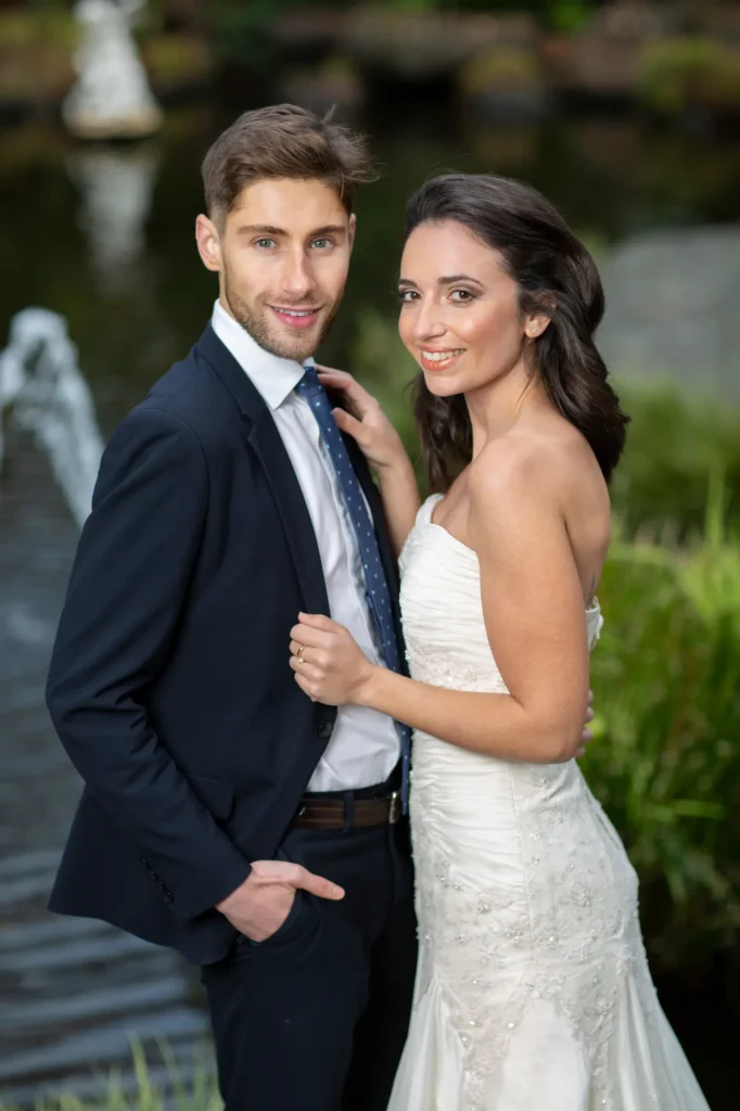 A bride and groom share an intimate moment in Fitzroy Garden, surrounded by lush greenery and a tranquil pond. The groom, dressed in a dark suit with a blue tie, stands confidently beside his bride, who wears a strapless white gown adorned with delicate beading. She smiles warmly, her hand gently resting on his chest as they pose together in soft natural light. The peaceful garden setting, complete with a fountain and vibrant foliage, enhances the romantic atmosphere of this garden wedding portrait. Captured in Melbourne, this image beautifully reflects the joy and elegance of a wedding day in a serene outdoor setting.