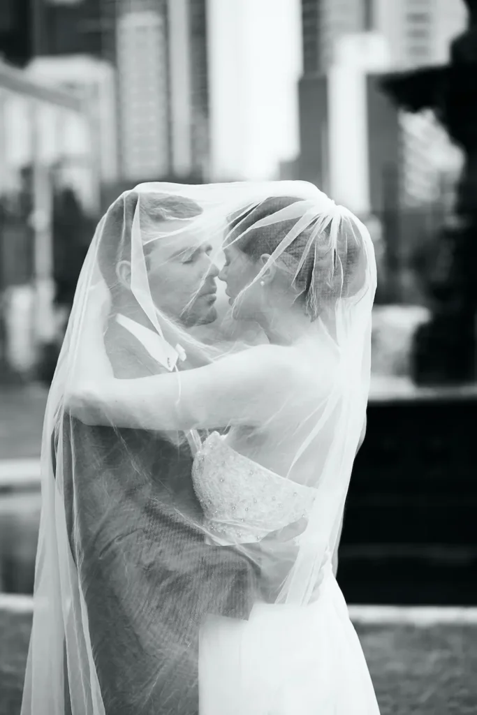 A romantic black-and-white wedding portrait of a bride and groom at the Golden Reserve in Melbourne. The couple embraces tenderly under the bride’s sheer veil, which drapes over both of them, creating an intimate and serene moment. The groom wears a dark suit with vest and tie, while the bride wears a strapless lace gown. A dark fountain in soft focus forms the backdrop, with blurred city buildings hinting at the urban park setting. The photograph highlights their emotional connection, capturing the timeless elegance and intimacy of Melbourne bride and groom portraits during a wedding celebration.