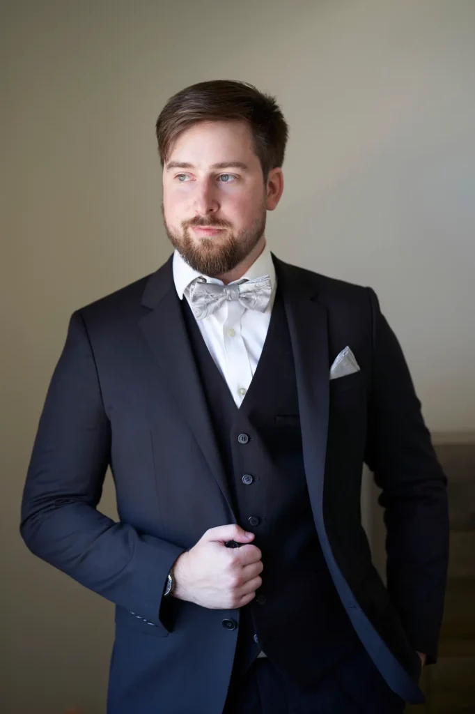 A refined indoor portrait capturing a quiet moment of reflection as the groom gets ready before the wedding. He stands slightly angled away from the camera, gazing thoughtfully to the left while adjusting the lapel of his dark suit jacket. The soft, natural light from the side highlights his face and shoulders, creating a gentle contrast against the neutral background. Dressed in a black suit with a white shirt, silver bow tie, and folded pocket square, he exudes calm confidence and timeless sophistication. The minimalist setting and subtle lighting emphasize the groom’s elegance and composure before the ceremony.