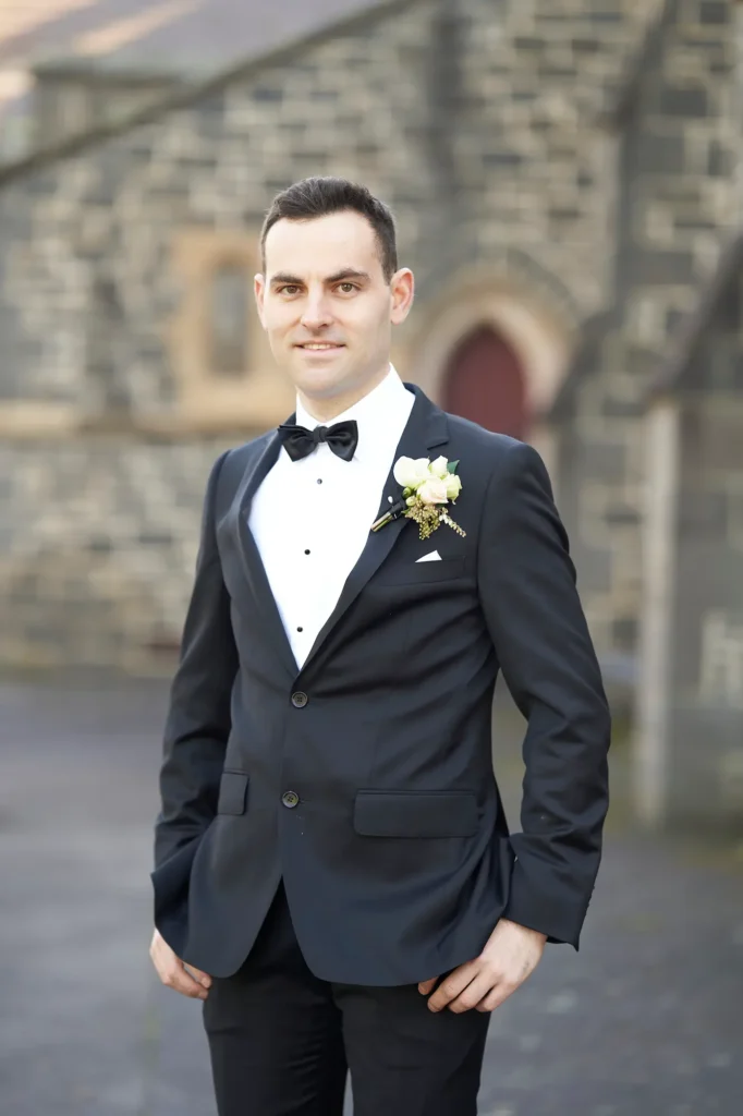 A groom stands outdoors in front of a historic stone building, his relaxed posture and warm smile capturing the quiet confidence of the moment. The soft, natural light highlights the texture of his suit and the subtle tones of the stone facade behind him. With his hands casually in his pockets and his body slightly angled, he faces the camera with an expression that feels both genuine and timeless. The blurred arches and muted colors of the background create a gentle contrast that draws focus to the groom, giving the portrait a refined and classic atmosphere.