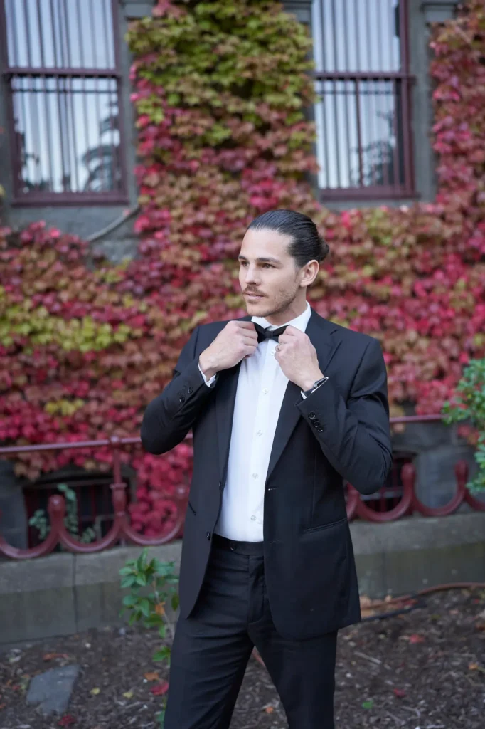 A groom in a classic black tuxedo adjusts his bow tie in front of the ivy-covered Victoria Barracks Museum in Melbourne. The historic stone building, adorned with autumn-colored vines, forms a rich and textured backdrop for this elegant wedding portrait. Sunlight softly illuminates the groom’s thoughtful expression, highlighting the crisp details of his attire and the warm hues of the ivy behind him. The composition balances timeless style with natural beauty, capturing a moment of quiet confidence and sophistication — a signature Melbourne wedding photography portrait set against the heritage charm of Victoria Barracks.