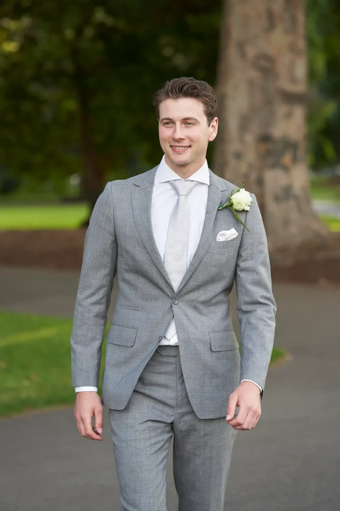 A joyful outdoor wedding portrait of the groom walking along a paved path in a lush green park. Captured mid-stride, he smiles warmly and looks to his left, radiating happiness and quiet confidence. The background features a tall, textured tree and soft greenery, creating a serene, natural setting. Dressed in a light gray suit with a white shirt, patterned tie, and white rose boutonnière, he embodies timeless elegance. The bright daylight enhances the cheerful mood and relaxed energy of the moment. A perfect example of candid and natural wedding photography in Melbourne, capturing genuine emotion and effortless style.
