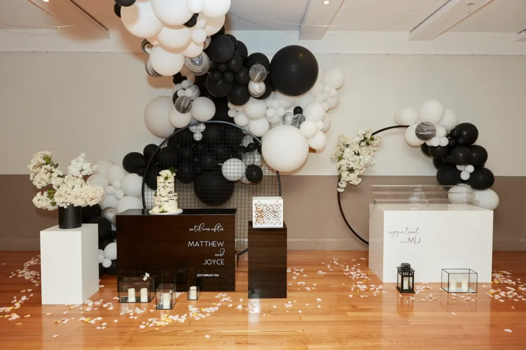 A sophisticated Melbourne engagement party setup featuring a chic multi-tiered white cake with black and white details and greenery, displayed on a dark console table with elegant script reading "MATTHEW AND JOYCE." Behind it, a dramatic black and white balloon arch surrounds a large black circular frame, complemented by fresh white floral arrangements in black vases. White petals and confetti scatter across the wooden floor, while candles and subtle lighting enhance the romantic, modern ambiance. Professional photography captures the stylish and celebratory atmosphere of this memorable engagement party.
