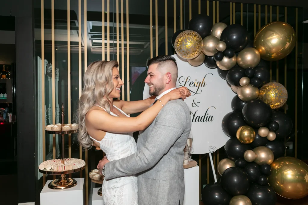 A joyful engaged couple shares a tender embrace at their Melbourne engagement party. The bride-to-be wears an elegant white halter-neck dress with glamorous waves, while the groom-to-be dons a grey suit jacket and white shirt. Behind them, a white circular sign displays "Leigh & Nadia," framed by a sophisticated black and gold balloon arch. A multi-tiered dessert stand with pastries adds to the festive atmosphere, captured beautifully in professional photography highlighting intimacy, happiness, and stylish party decor.