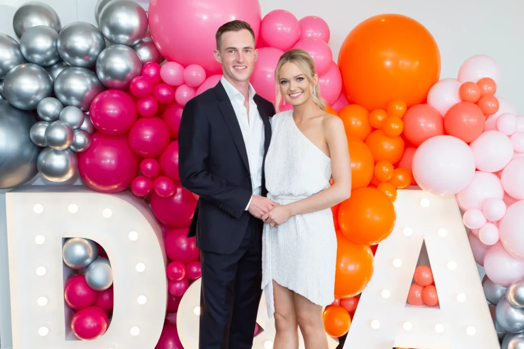 svg%3E A joyful engaged couple celebrates their engagement in front of a colorful balloon backdrop featuring pink, silver, and orange balloons and large illuminated letters "DA". The bride-to-be shines in an elegant white one-shoulder sequin mini dress, while the groom-to-be looks dapper in a dark suit with a white shirt. They smile and pose together, capturing a lively, festive, and personalized moment of their engagement party, surrounded by vibrant decorations that reflect their happiness and the celebratory atmosphere of the occasion.