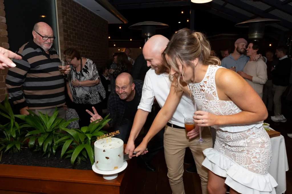 The engaged couple joyfully cuts their white two-tiered cake at a Melbourne engagement party. The bride-to-be wears a sleeveless white lace mini-dress with a high-neck and ruffled hem, while the groom-to-be dons a white shirt and light-colored trousers. The cake features gold accents and a "Mr & Mrs" topper, set on a table with lush greenery. Friends and family gather around, smiling and celebrating in a warmly lit venue. Professional photography captures this central, happy, and festive moment of the engagement party.