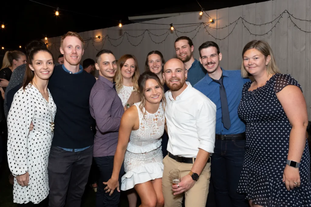 A joyful engaged couple celebrates their engagement party in Melbourne surrounded by friends and family. The bride-to-be wears a chic white lace mini-dress with a high neckline and ruffled hem, and the groom-to-be stands beside her in a white shirt and tan trousers, holding a drink. Guests in smart-casual attire smile and interact around them. Strung lights illuminate the fenced area, creating a warm, festive, and intimate atmosphere, captured beautifully in professional photography that highlights camaraderie and shared happiness.
