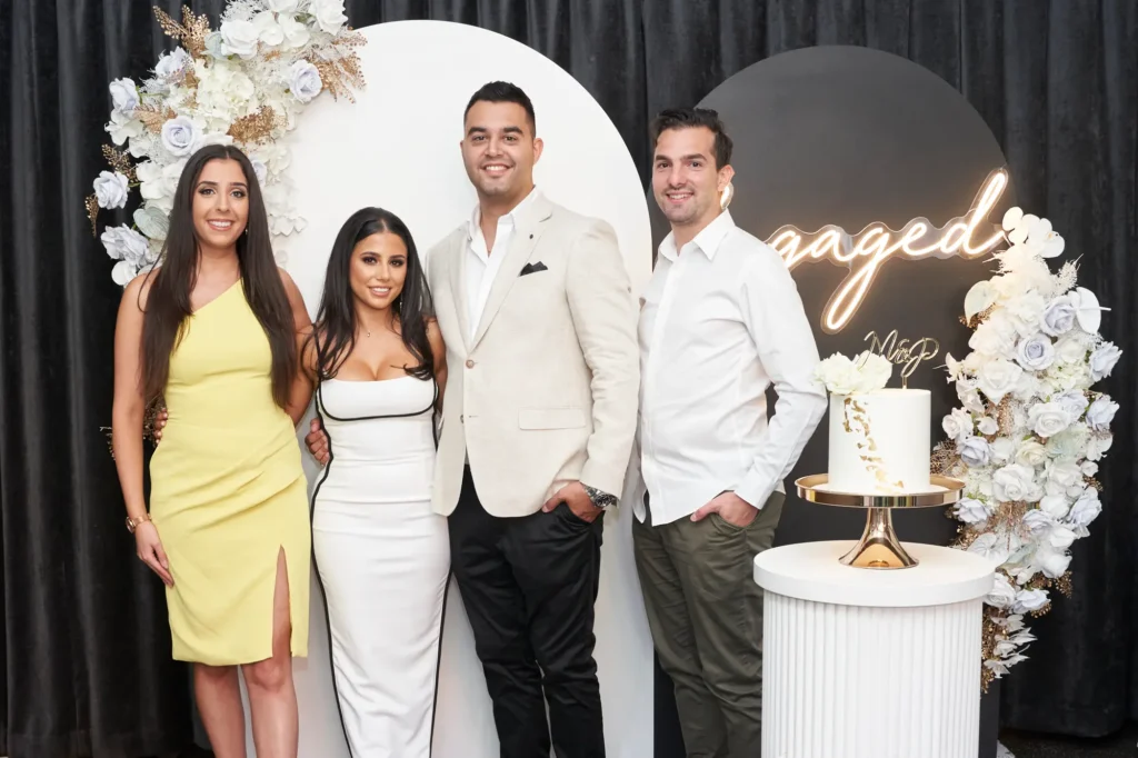 A joyful engaged couple celebrates their Melbourne engagement party with two friends or family members. The bride-to-be wears a white midi dress with black piping and a sweetheart neckline, while the groom-to-be dons a light-colored jacket and dark trousers. Guests include a woman in a yellow one-shoulder dress and a man in a white shirt with olive trousers. Behind them, circular floral backdrops in white and gold, a gold neon "engaged" sign, and a decorated white tiered cake create a sophisticated, festive atmosphere, captured beautifully in professional photography.